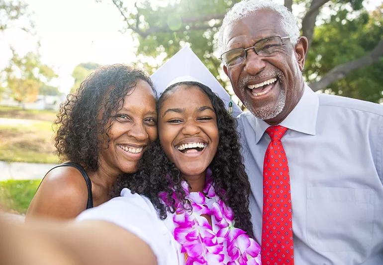 A young graduate smiles while taking a selfie next to their parents.