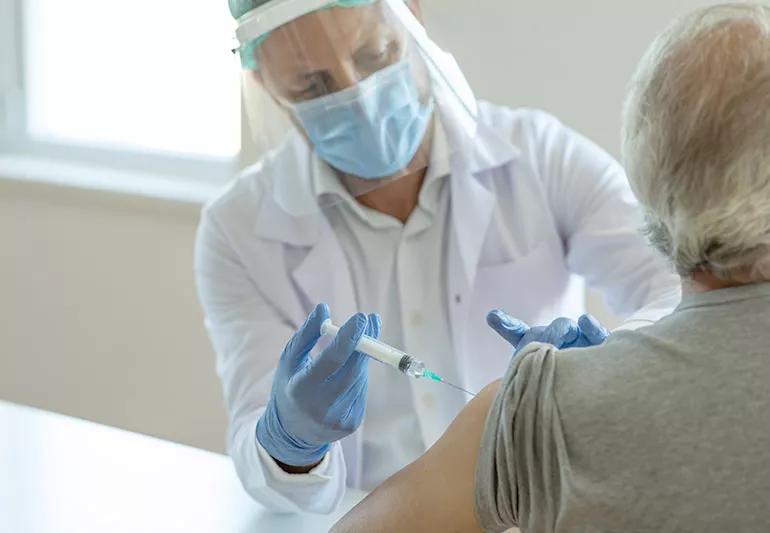 A healthcare provider wearing a face shield and a mask giving a shot in someone's left arm