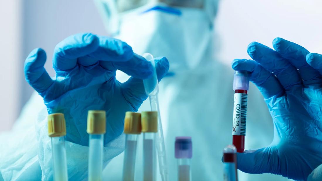 Person in mask and medical coveralls handling COVID-19 vials with gloved hands