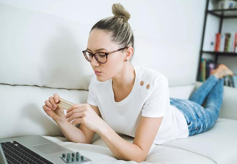 Woman lying a couch and looking at a packet of pills atop a laptop