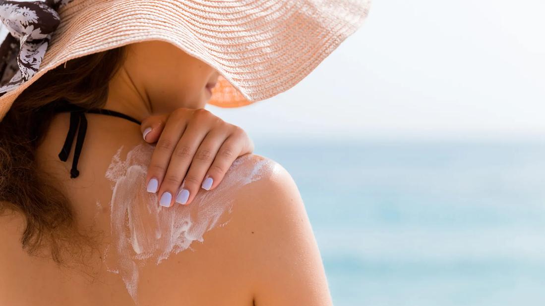 Person wearing big floppy hat on the beach, applying sunscreen to their shoulder