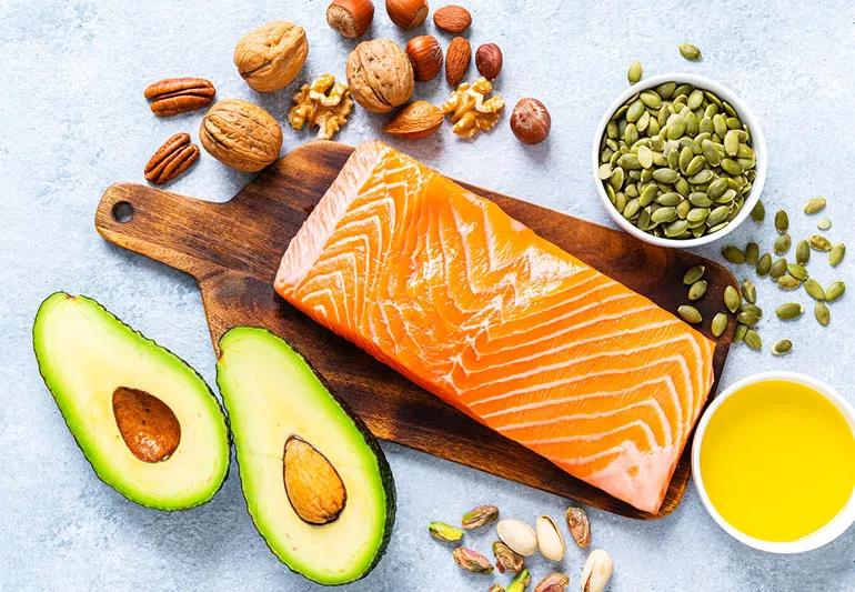 A piece of salmon sits on a cutting board surrounded by avocados, olive oil and various nuts.