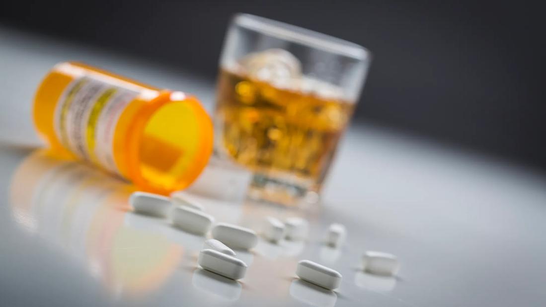 Open medicine bottle on its side, with spilled pills on the table, and glass of alcohol nearby