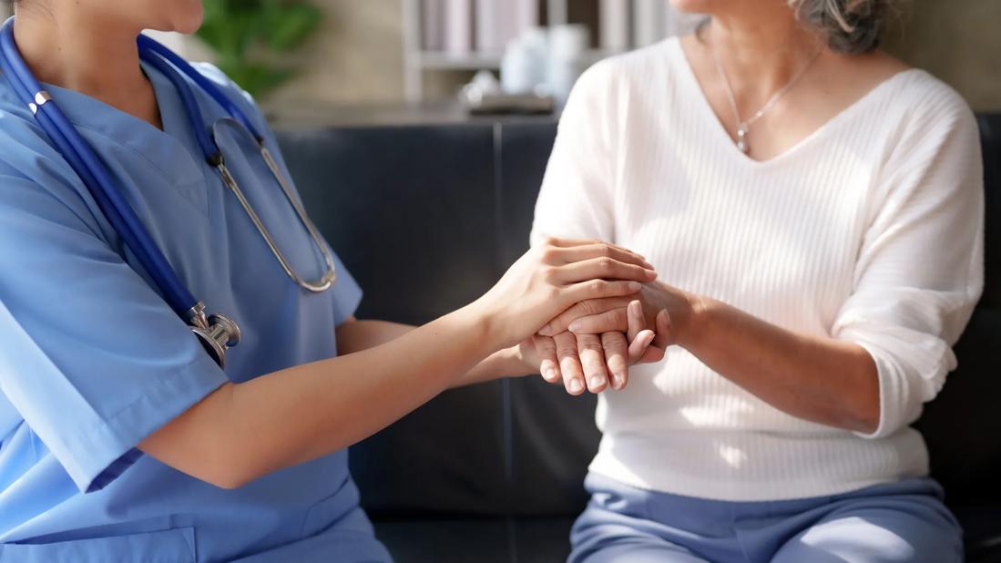 Healthcare provider in scrubs, stethoscope around neck, sitting and holding the hands of sitting person