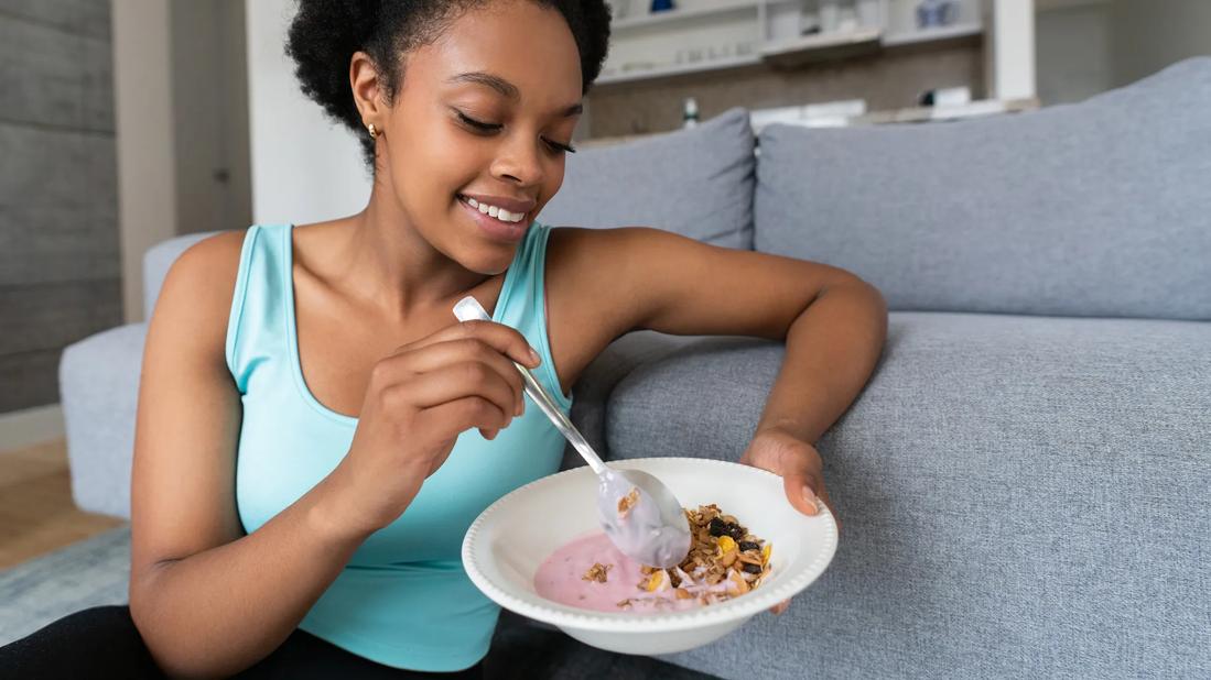 Smiling woman sitting on couch holding bowl of yogurt and granola