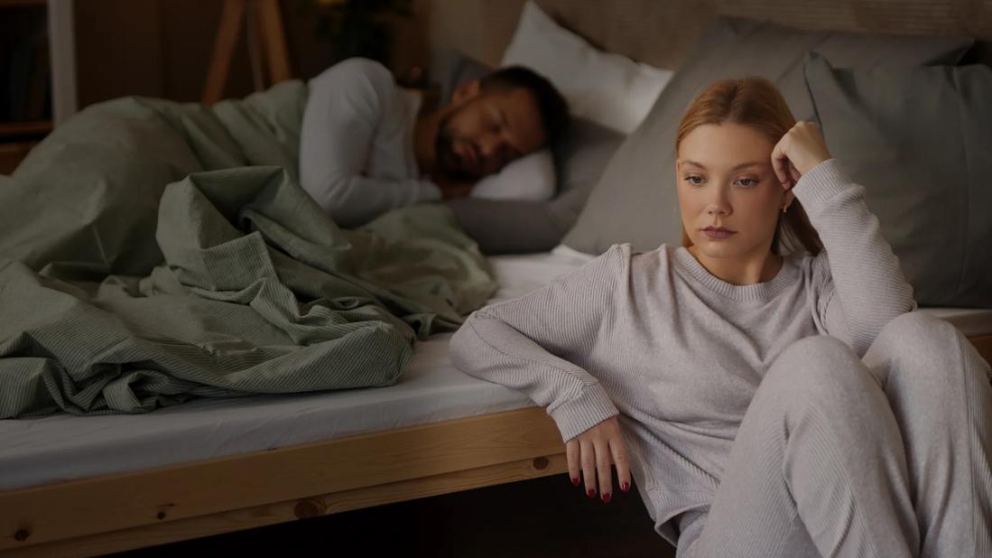 Person sitting on floor at night next to bed in deep thought, with partner sleeping in bed