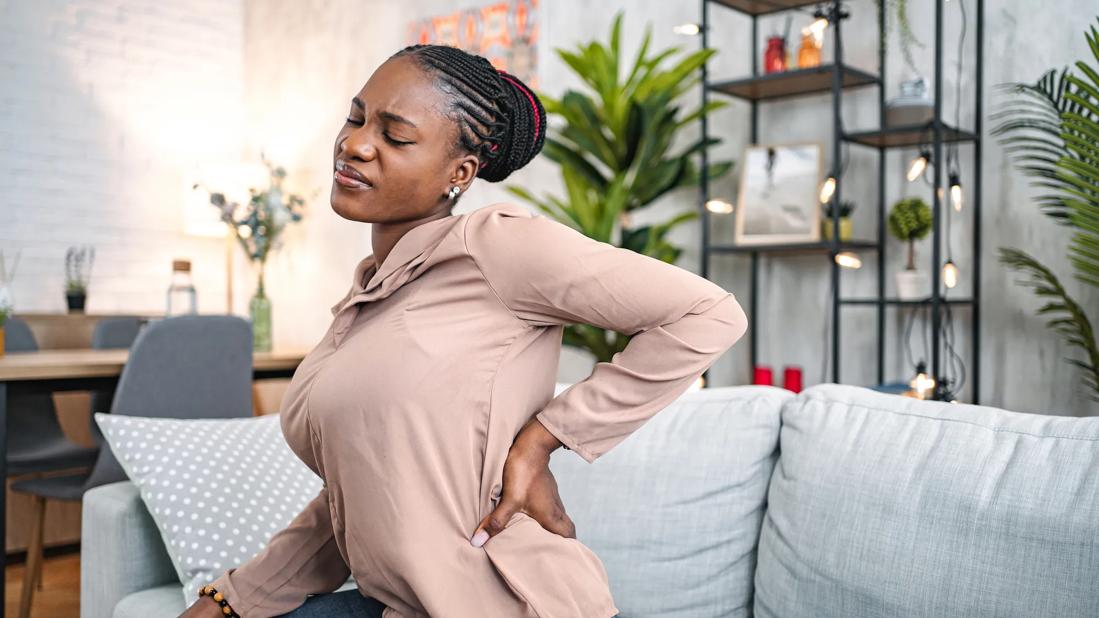 Person sitting on couch at home, with hand on lower back, grimacing