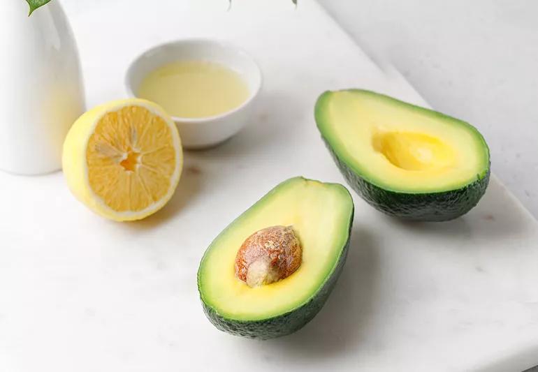 An avocado sliced in half with pit showing in one half, positioned on white marble with a sliced lemon in the background.