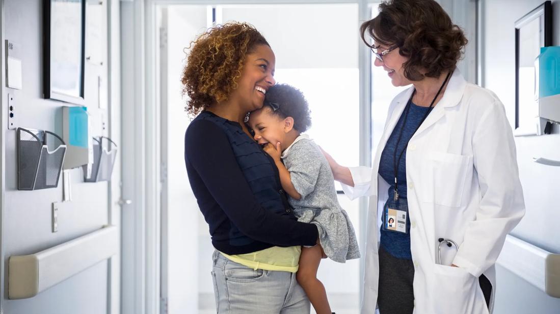 Smiling caregiver holding smiling toddler interacting with smiling healthcare provider in medical hallway