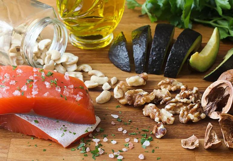 A cutting board with sliced avocados, olive oil, nuts, salmon and leafy greens