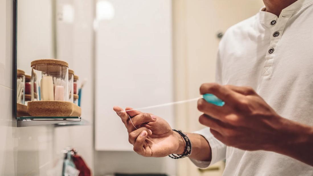 Person in bathroom, with jars of products on shelf, wrapping long piece of floss around fingers