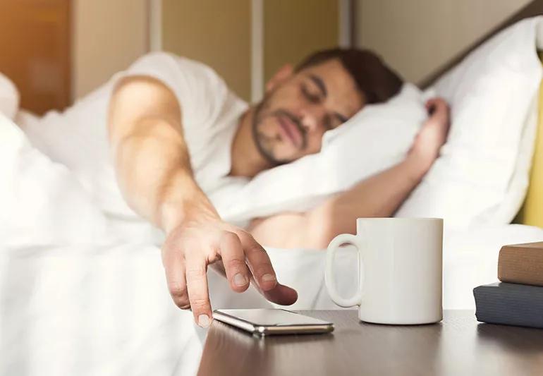 A person in bed with their eyes closed reaches for their smartphone