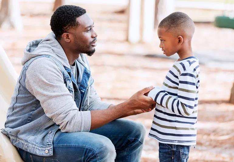 Man sitting across a young boy, talking and holding his hands in comfort.