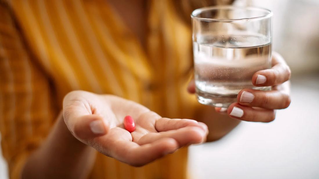 Hand holding red capsule, with other hand holding glass of water