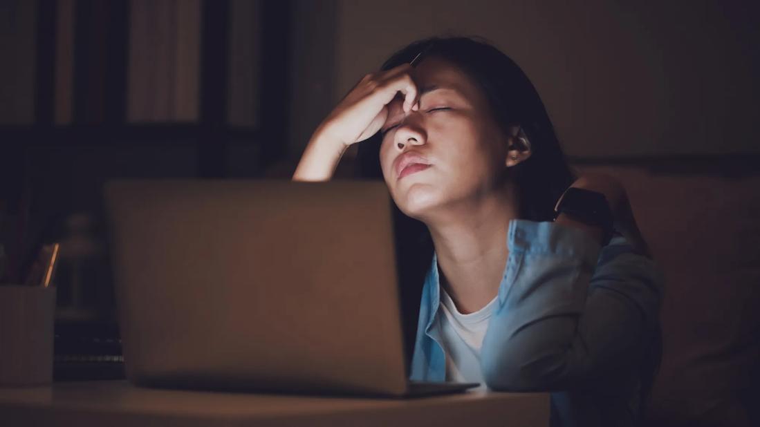 Person in front of their laptop, pinching their forhead, eyes closed, room dark, laptop light shining