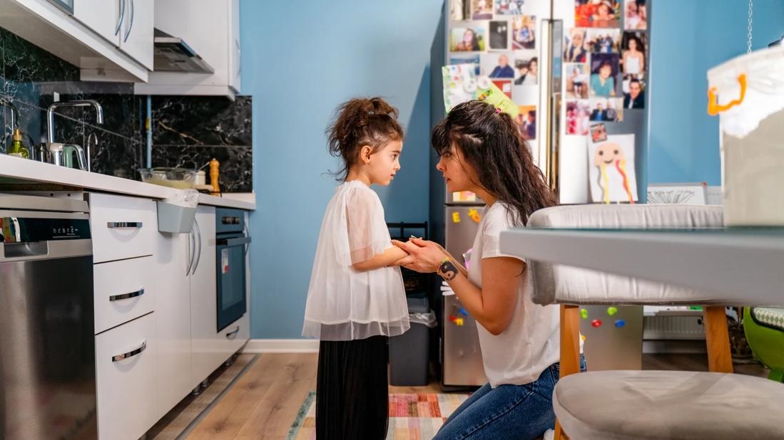 Caregiver kneeling down at home, holding child's hands, with stern look on caregiver's face