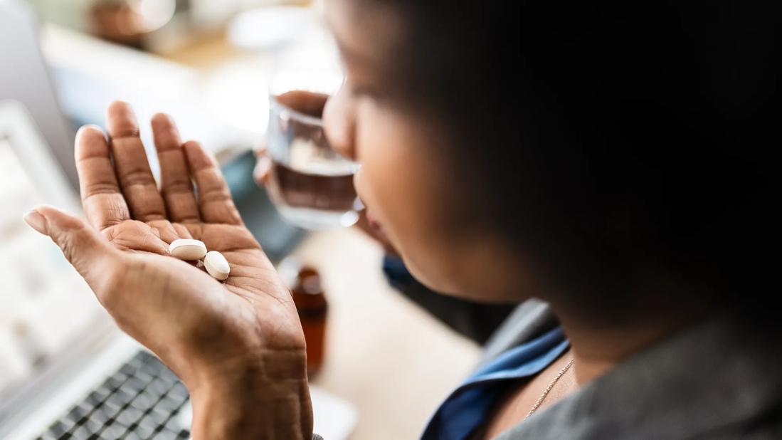 Person holding two pills in palm of hand and glass of water in other hand, keyboard in background