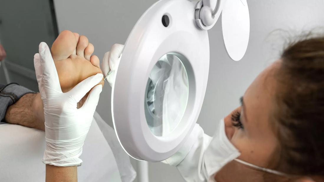 Medical technician looking through large, lighted magnifying glass, working on patient's foot