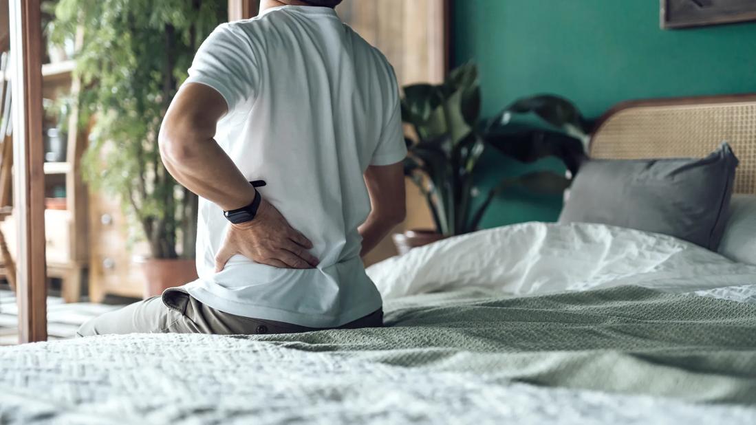 Person sitting on edge of bed with hand on lower back, arched in discomfort