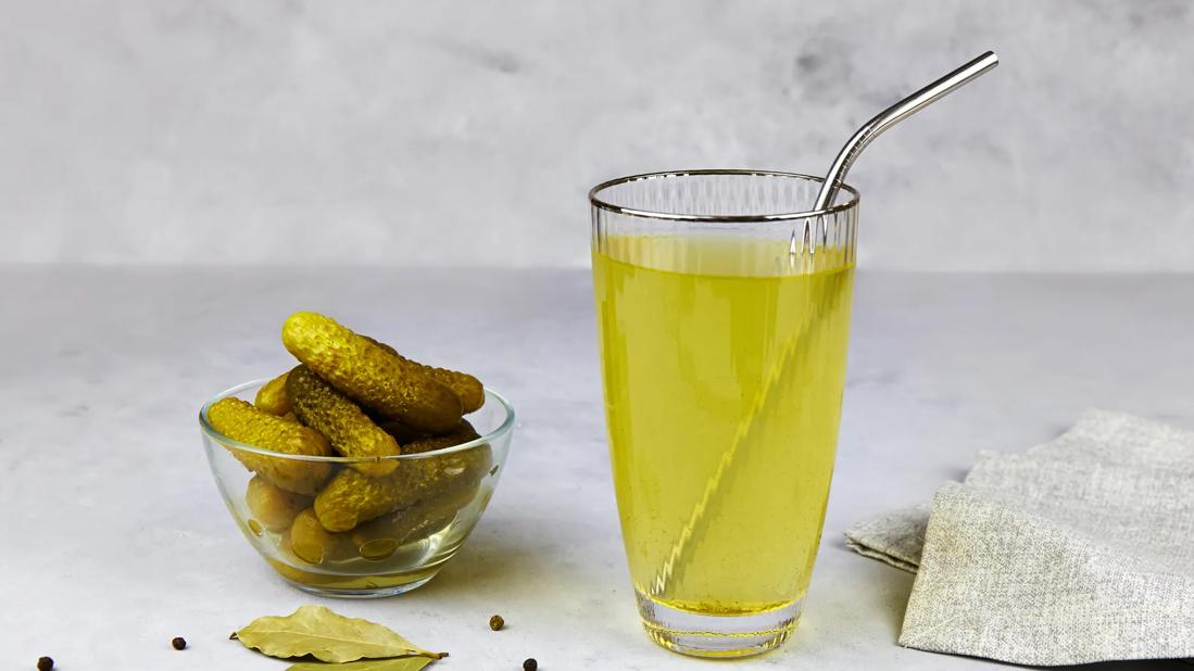 Glass of pickle juice with metal straw and glass bowl of pickles