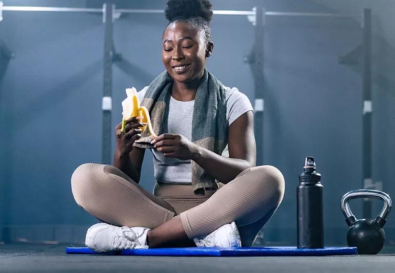 Person eating banana after exercising while sitting cross-legged on floor mat with towel around neck.