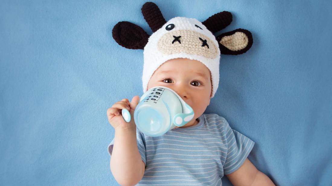 Baby lying down with crocheted cow hat on, drinking from sippy cup