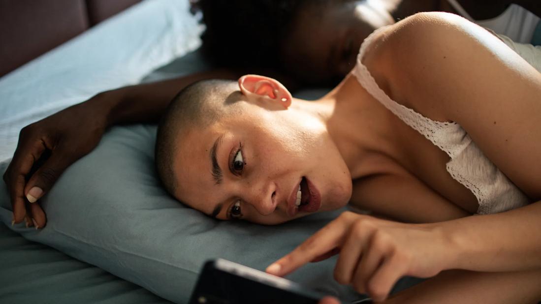 Couple in bed, one asleep, the other typing on their cell phone