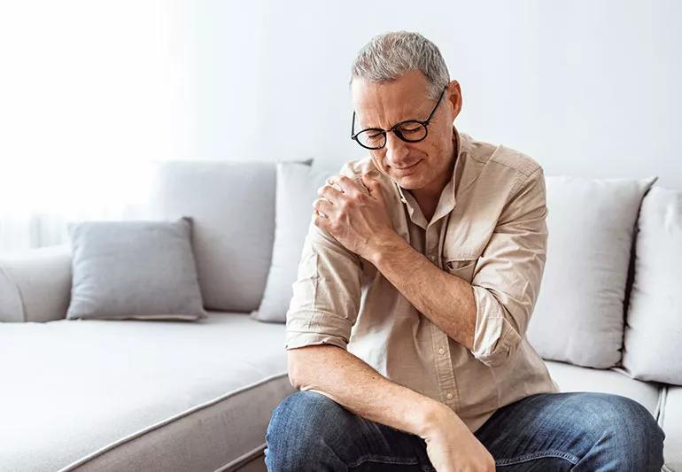 A man with silver hair and glasses holding his right shoulder wile grimacing in pain