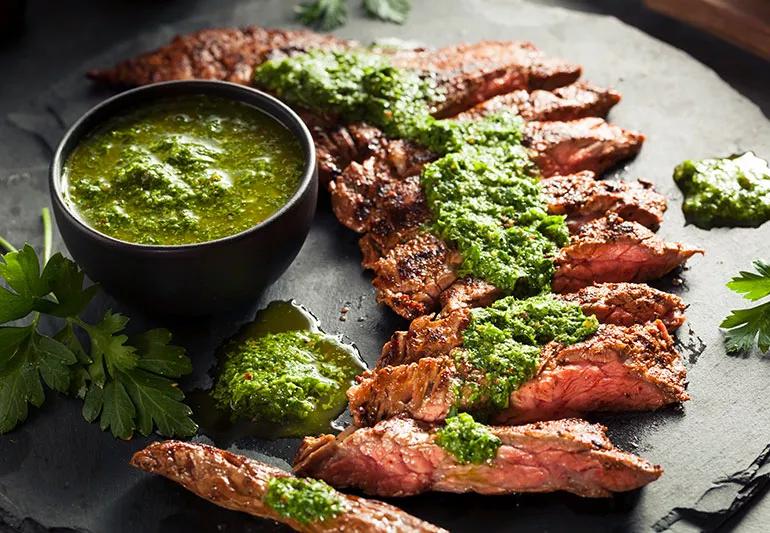Cuts of flank stake covered in an herbaceous marinade arranged around a bowl of extra marinade