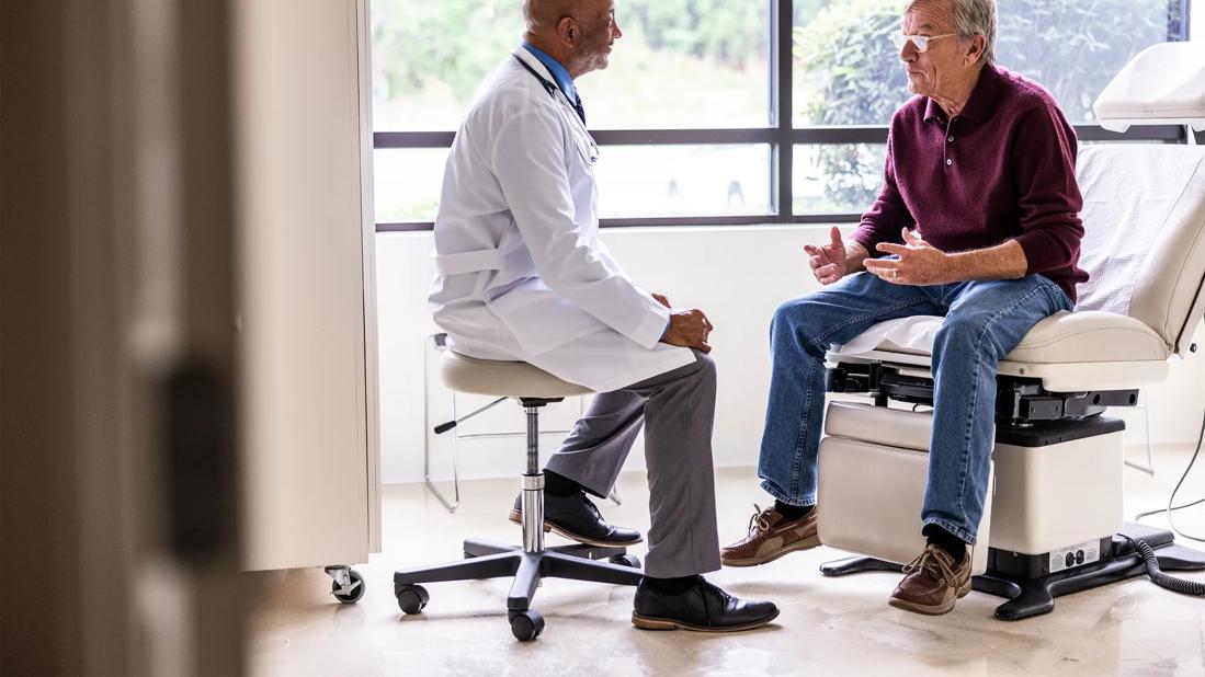 Older male in doctor's office sitting on exam chair, talking with healthcare provider