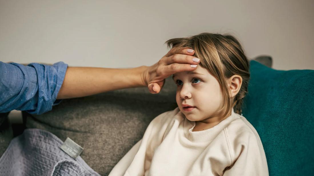 Hand feeling the temperature on the forehead of a toddler sitting on a couch