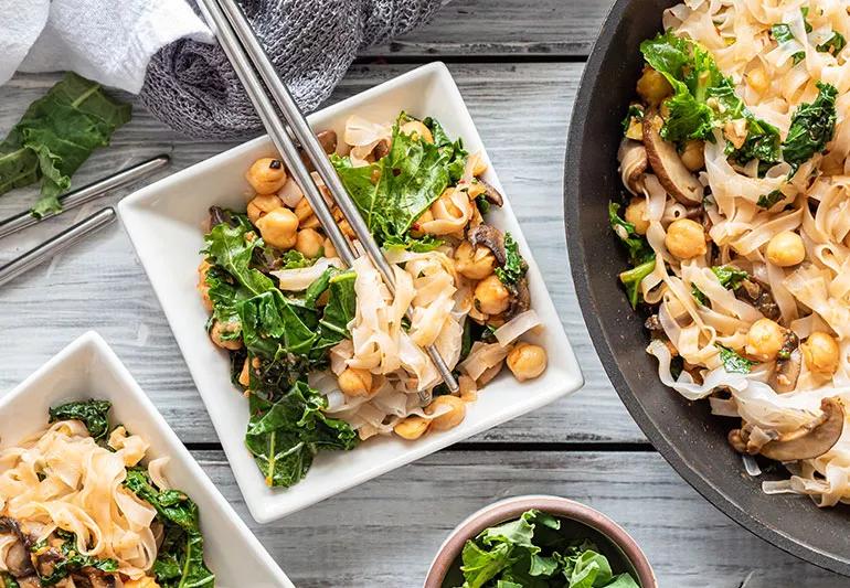 Silver chopsticks nestled in a bowl of noodles mixed with kale and chickpeas