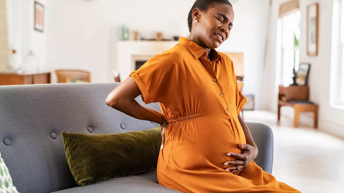 Pregnant woman sitting on couch at home holding her stomach and back, wincing in discomfort