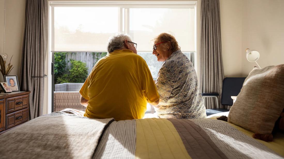 Elderly couple sitting on bed talking in bedroom, in early morning light