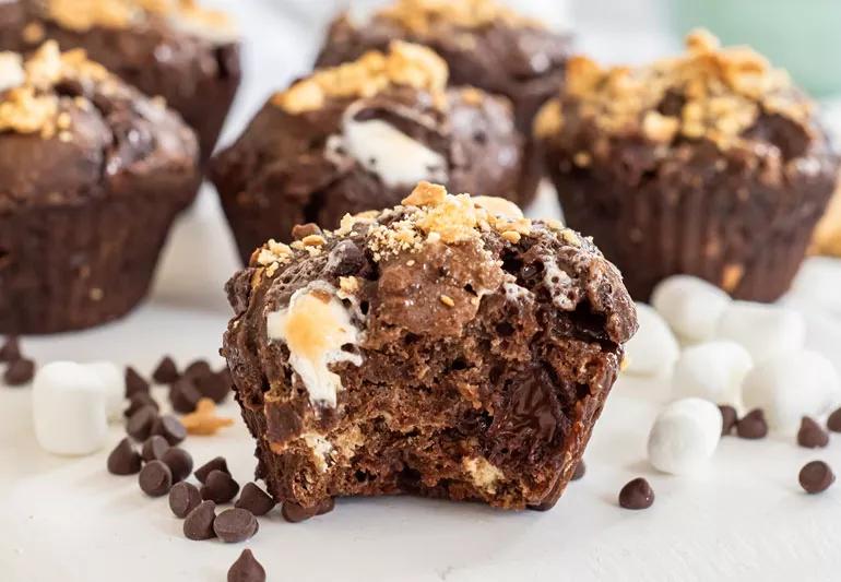 Closeup of S'mores muffins sith a bite taken out of one, with marshmallows and chocolate chips in the foreground.