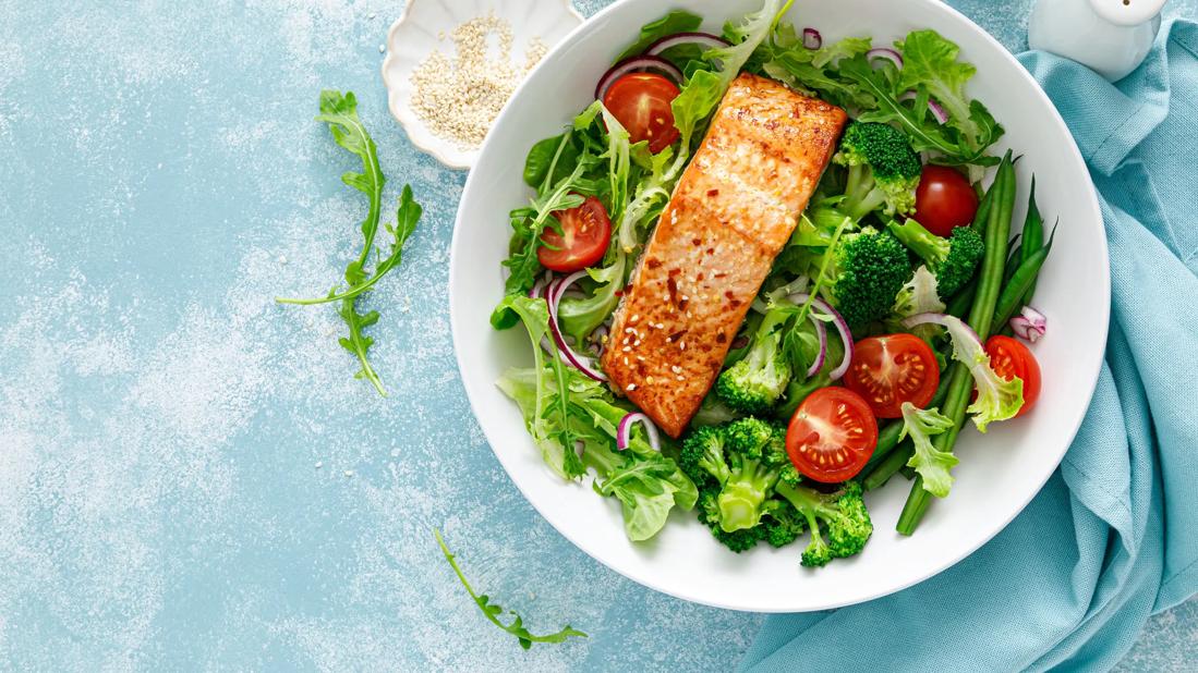 Plate filled with mixed greens and veggies and grilled salmon, with small bowl of sesame seeds and a slice of lemon on table