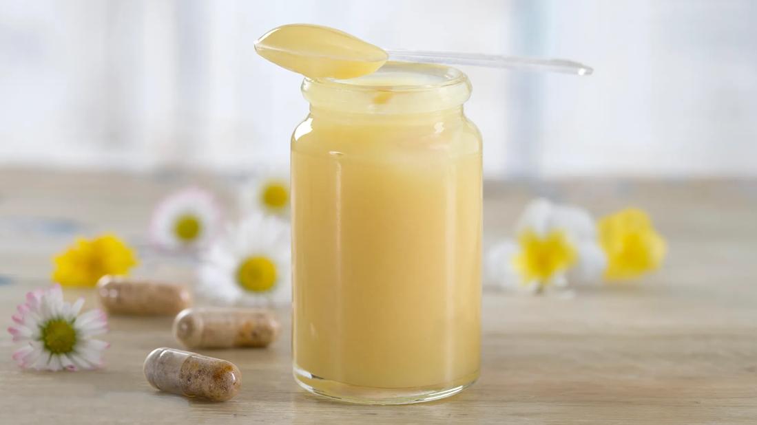 Jar of royal jelly with spoon on top, with capsules and flowers on table