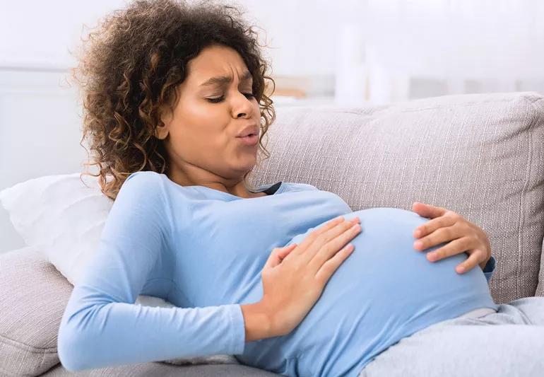 Pregnant woman sitting on couch at home holding her stomach and back, wincing in discomfort