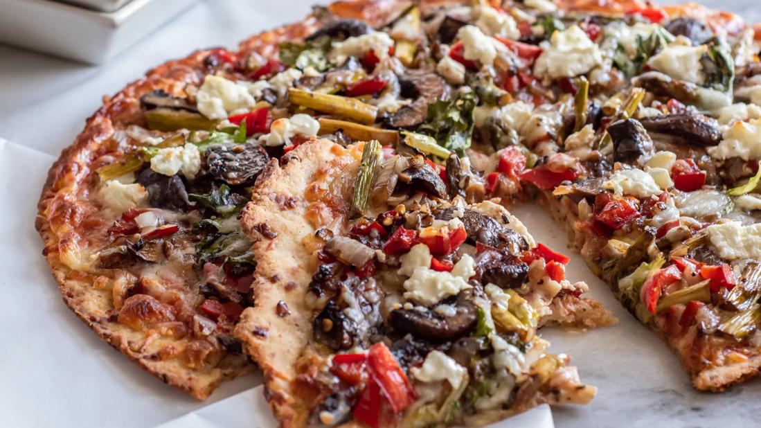 Whole arugula, veggie and goat cheese pizza, with one piece cut, with asparagus in background