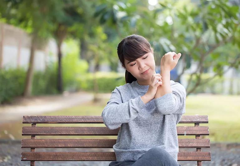 person in sweatshirt sitting on a bench outside and scratching their arm