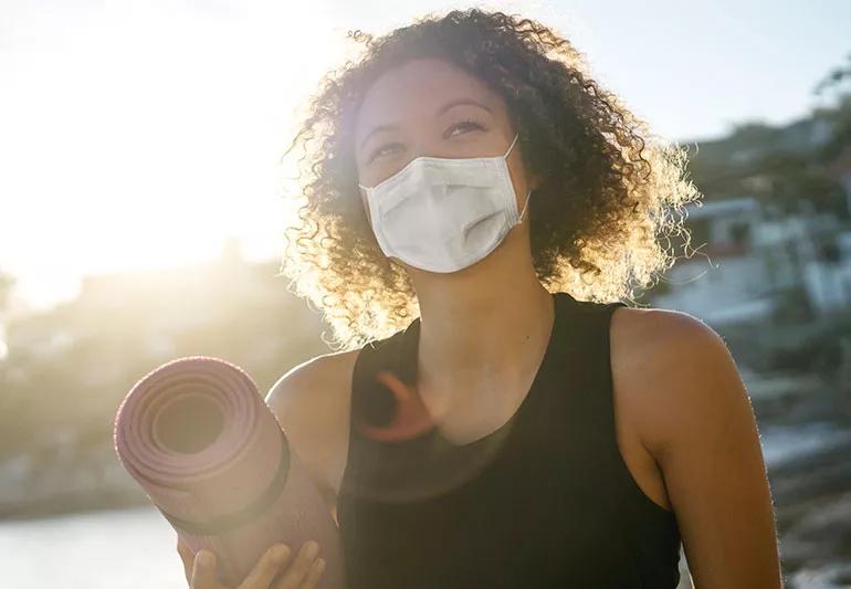 A curly-haired person with a surgical face mask wearing a black tank top and holding a yoga mat