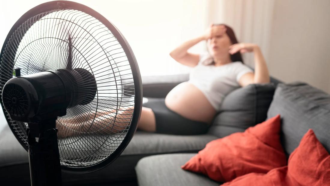 Fan blowing on pregnant person reclining on couch, with hand on forehead