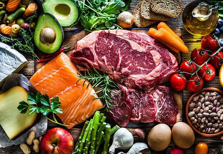 An assortment of food is displayed on a table, including raw steak, salmon, avocados, eggs, garlic, tomatoes and more.