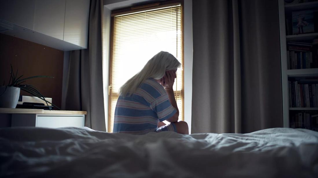 Person sitting on bed in pjs with head in hand, eyes closed