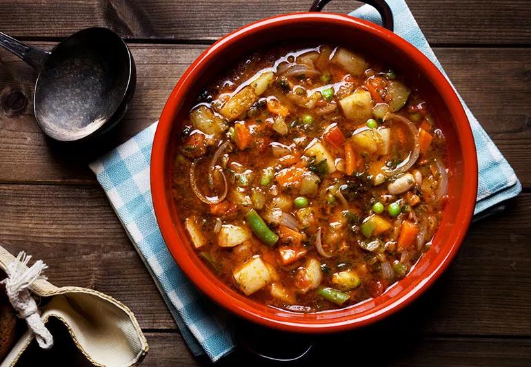 Red bowl filled with hearty minestrone soup sits atop a blue plaid napkin on a wooden table