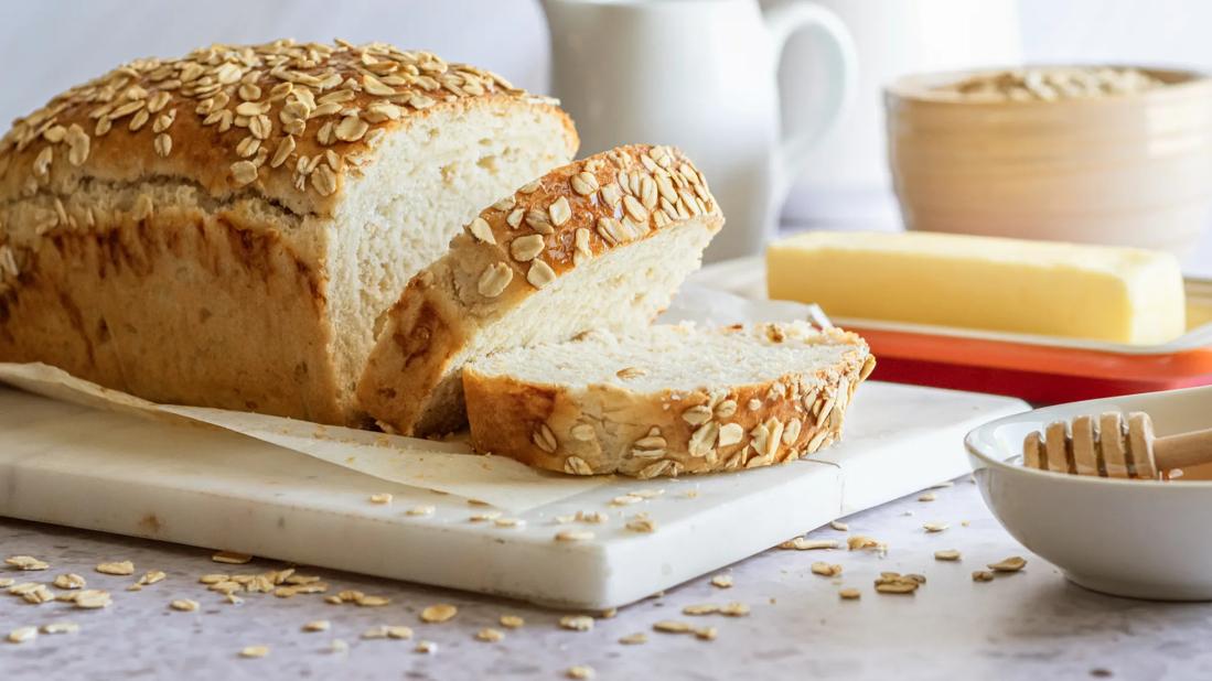 Partially sliced loaf of oats and honey bread on cutting board, with honey, butter nearby, oats sprinkled on table