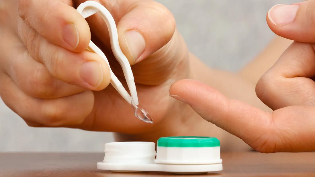 Hand holding plastic contact lens tweezer with contact in it, with contact lens container on table