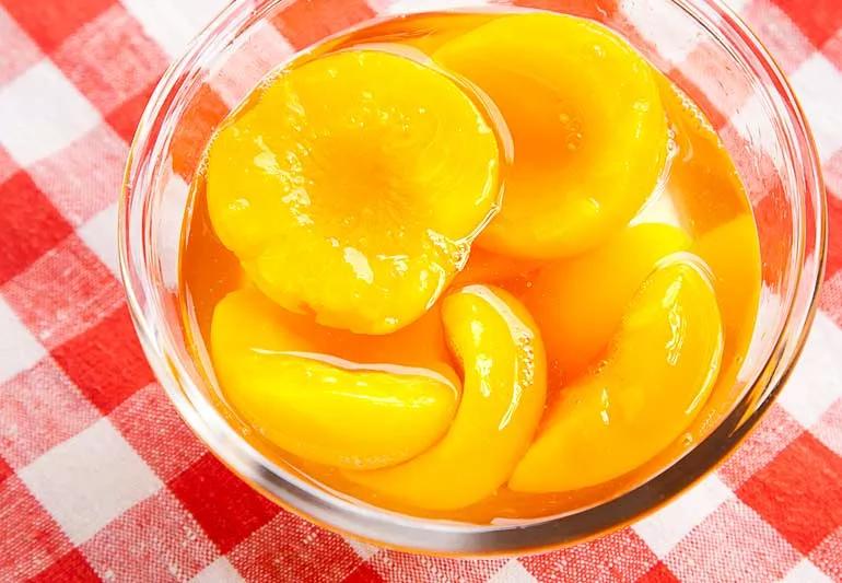 Peaches from a can are swimming in a sugary syrup in a bowl placed on a red checked tablecloth.