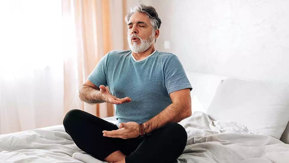 male doing yoga breathing exercises seated on a bed