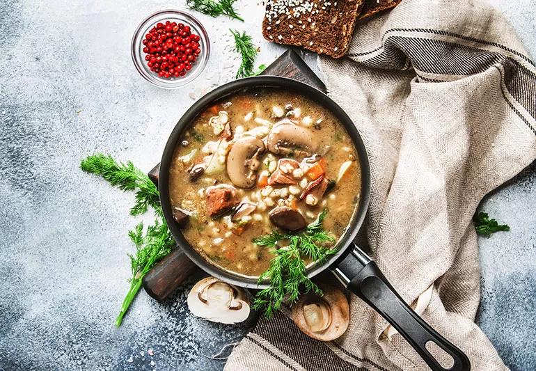 Pot of 3-mushroom barley soup on a cutting board surrounded by bread, berries, and a dish towel